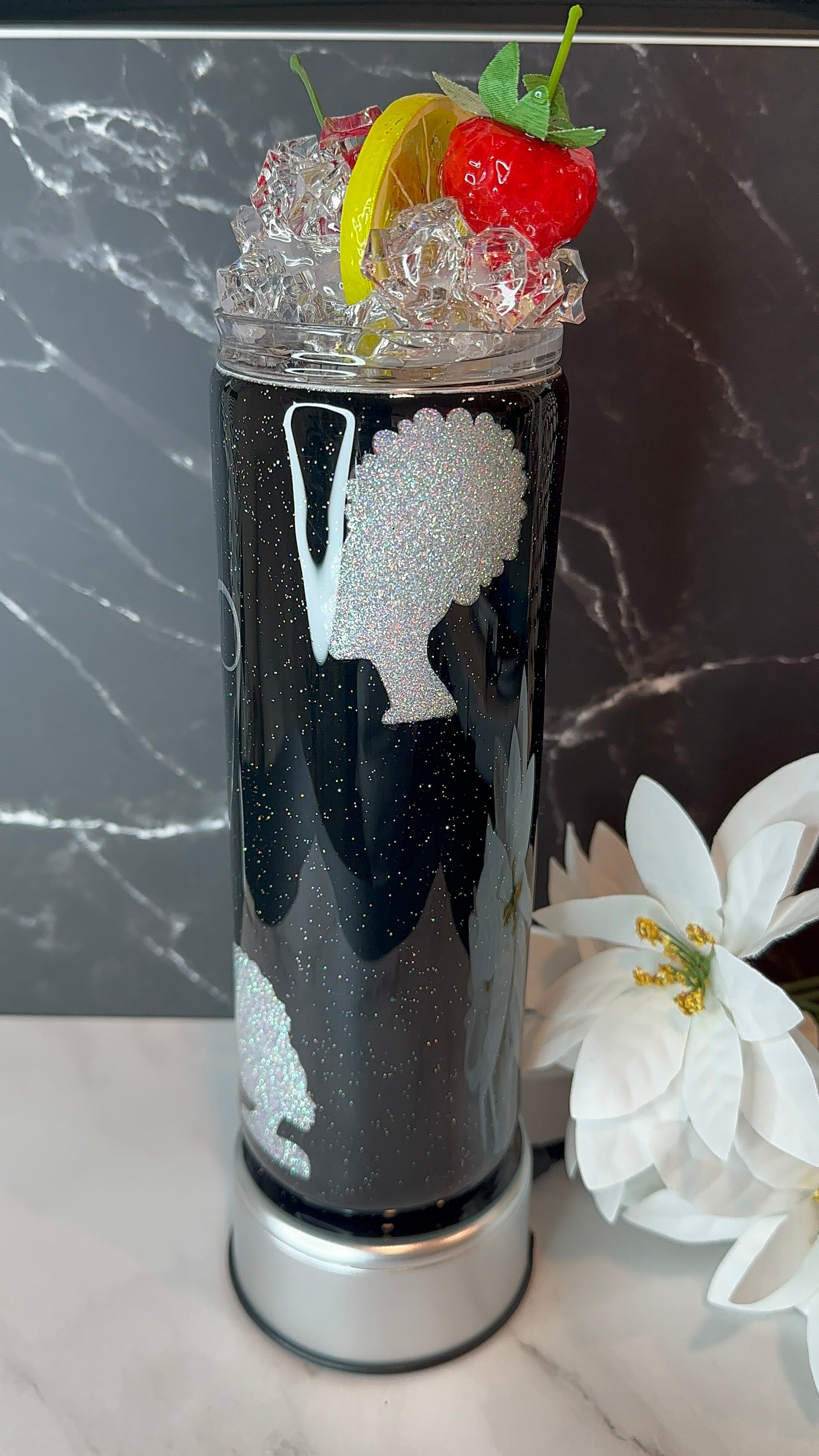 Tall black cylindrical container with white design, filled with ice and garnished with fruit, on a marble surface.