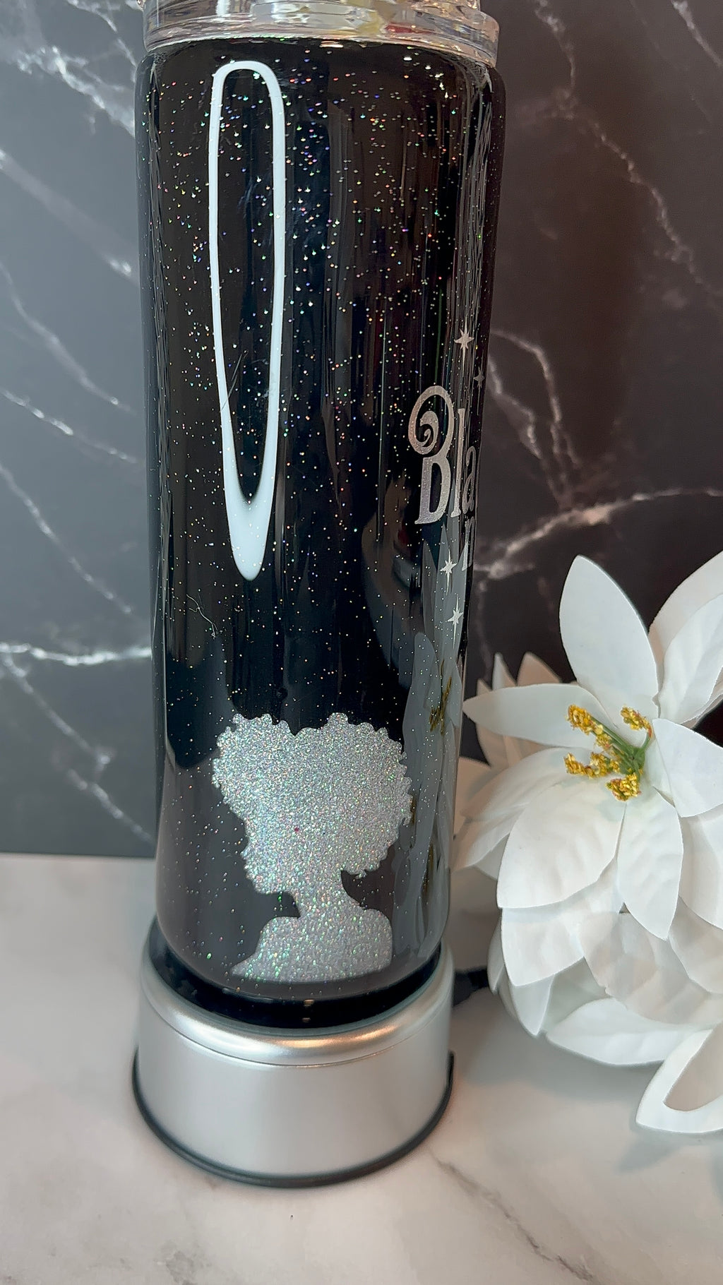 Black glittery tumbler with a logo on a marble surface with flowers.