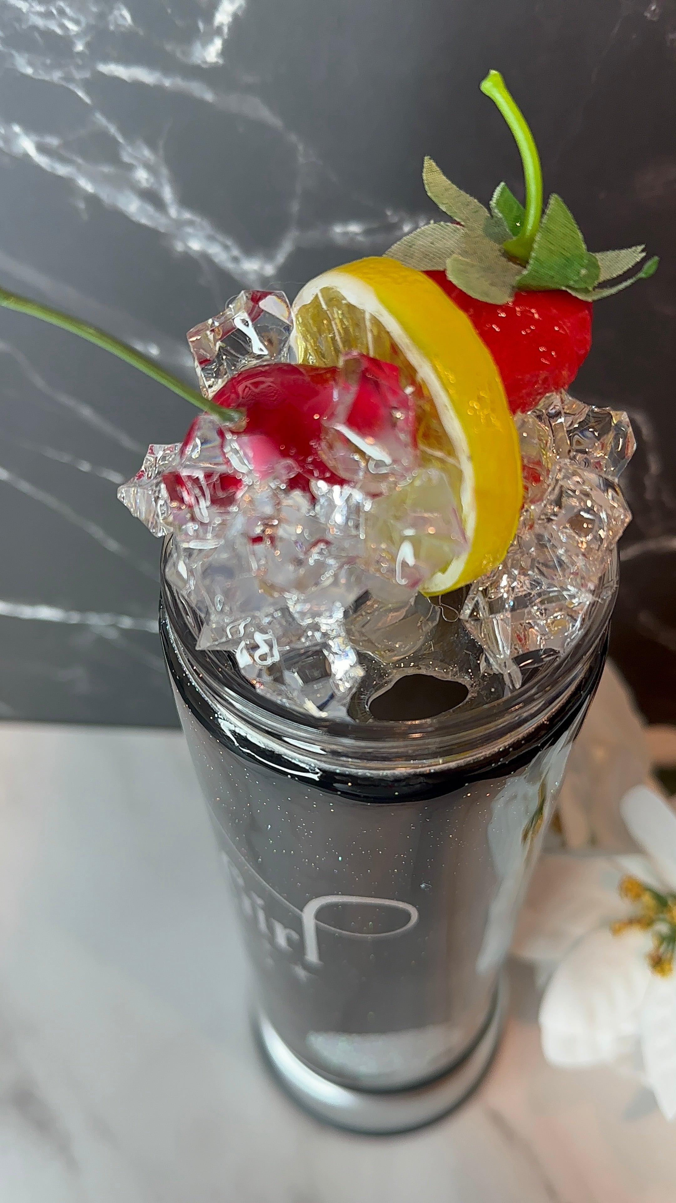 Stainless steel tumbler with a cocktail garnished with lemon, cherry, and ice cubes on a marble surface.