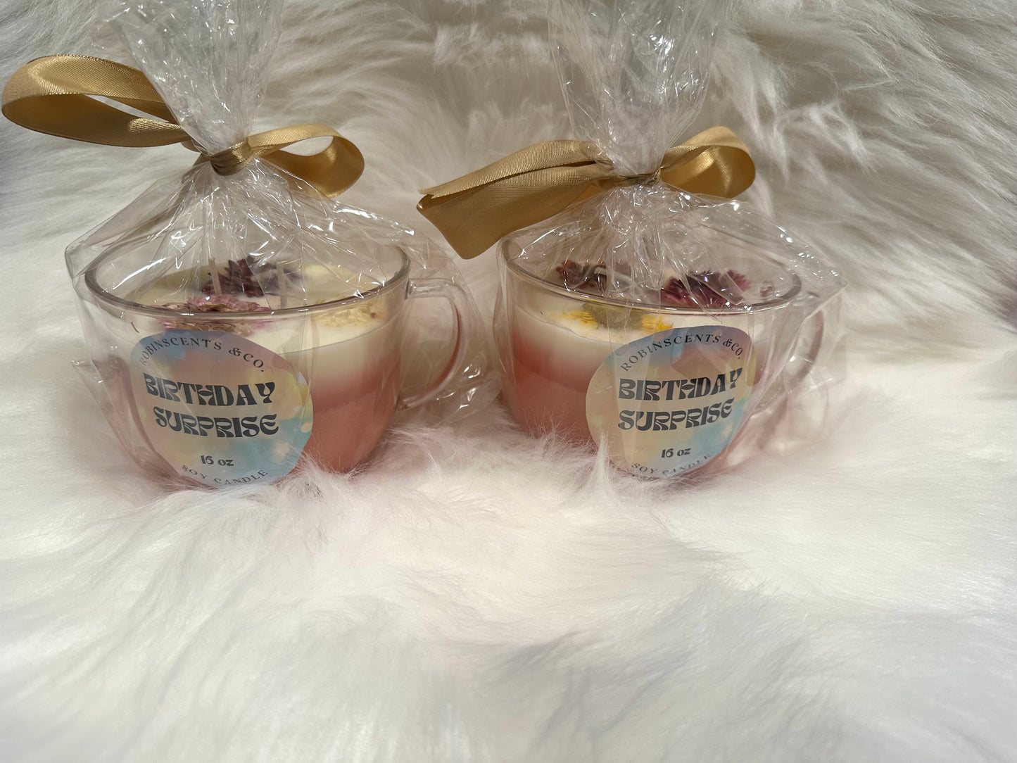 Two small glass jars with a 'Birthday Surprise' label, wrapped in clear plastic with gold ribbons on a white fluffy background.
