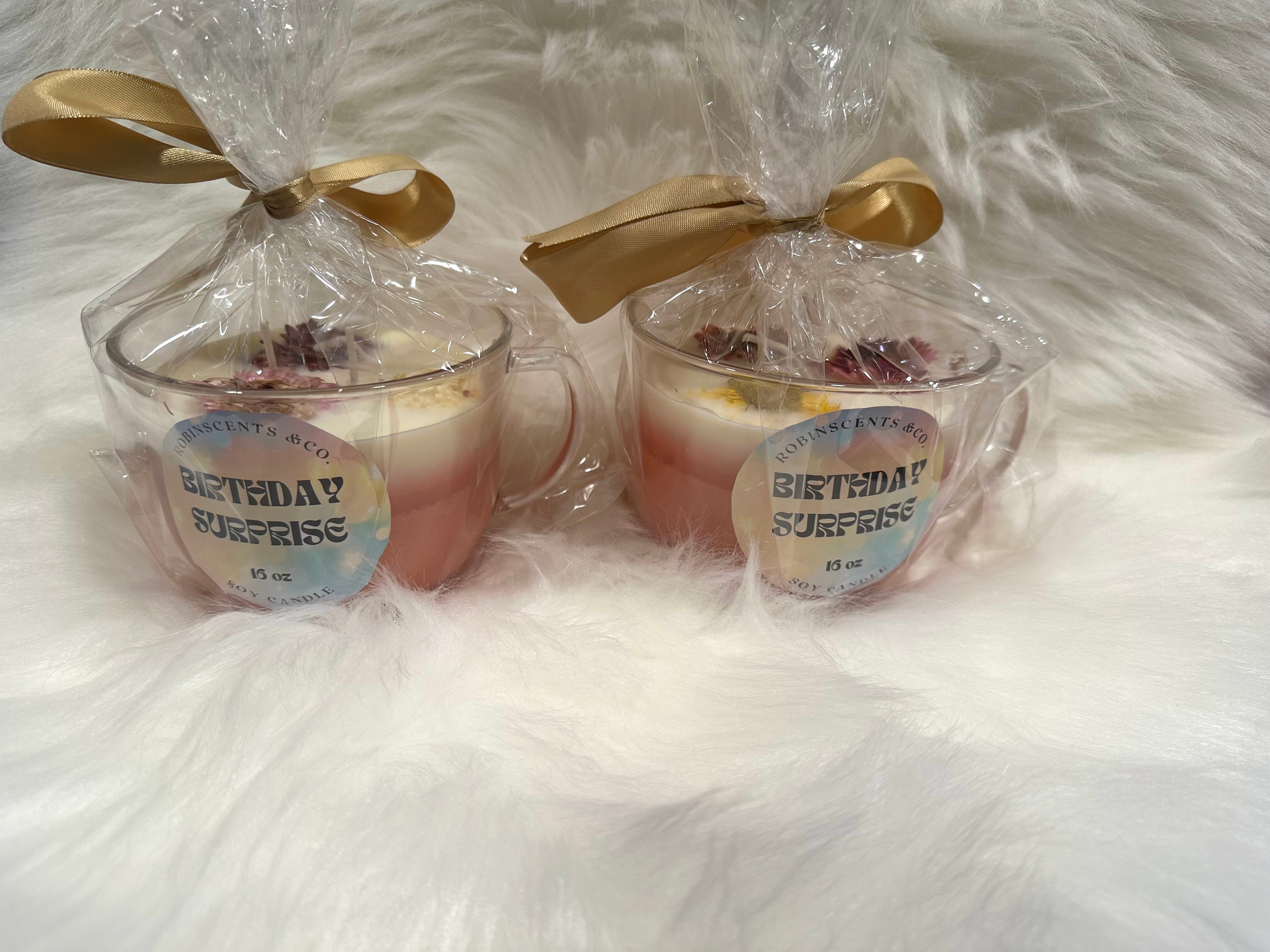 Two small glass jars with a 'Birthday Surprise' label, wrapped in clear plastic with gold ribbons on a white fluffy background.