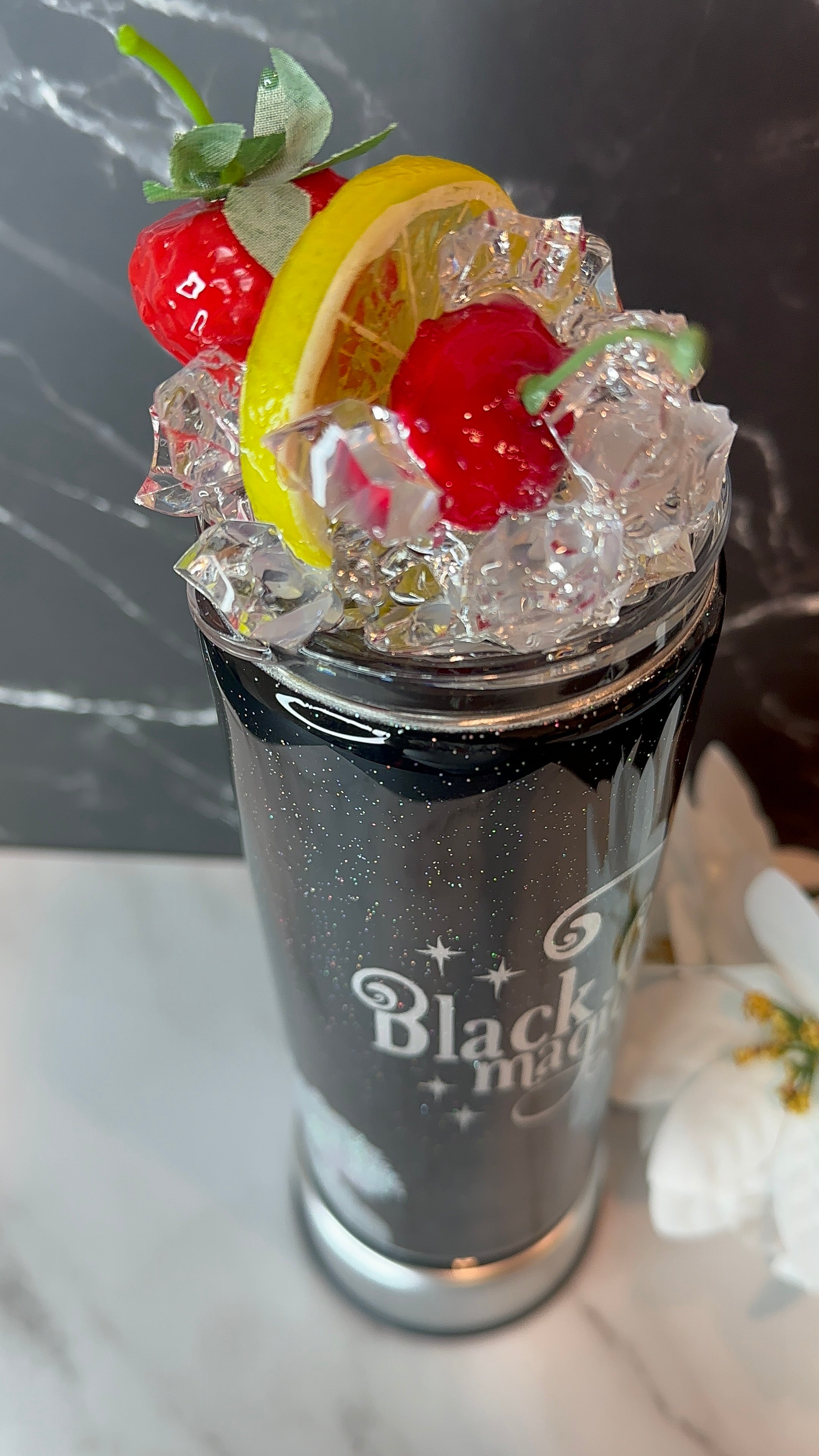 Decorative cocktail with fruit and ice in a glass labeled 'Black Magic' on a marble surface.